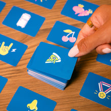 Load image into Gallery viewer, Hand playing a card game with blue cards featuring colorful illustrations from the board game Tilt n Shout on a wooden table.