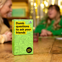 Load image into Gallery viewer, Green game box titled &#39;Dumb questions to ask your friends&#39; on a wooden table with blurred people in the background.
