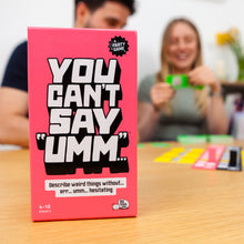 Load image into Gallery viewer, Pink game box titled &#39;You Can&#39;t Say Umm&#39; on a table with people playing and laughing in the background.
