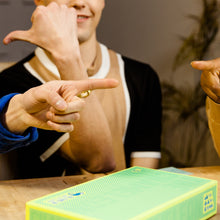 Load image into Gallery viewer, People pointing at the Chameleon with a green and yellow box on the table
