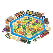 Load image into Gallery viewer, A board game set arranged on a white background, featuring a large hexagonal game board with a blue ocean border and a sandy island layout, surrounded by game boxes, cards, tokens, dice, and other game components. The word 'CATAN' is visible on several boxes.
