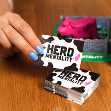 Load image into Gallery viewer, Hand holding a card with &#39;Herd Mentality&#39; design on a wooden surface
