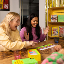 Load image into Gallery viewer, Two women playing the board game Dumb Questions To Ask Your Friends together at a table with books and snacks in the background.
