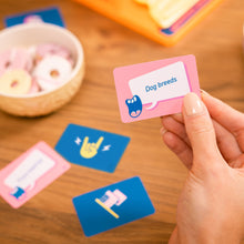 Load image into Gallery viewer, Hand holding a pink card from the board game Tilt n Shout  with &#39;Dog breeds&#39; text, surrounded by other cards on a wooden surface.
